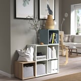 Modern living room with EKET shelf unit styled against art, holding books, a bag, vase, and decor near a cozy sofa and chair.