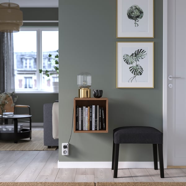 Wooden EKET wall cabinet with books, between art prints, gray stool below.