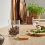 Small EKLATANT jar with bamboo lid, filled with dried mushrooms. On a kitchen table near herbs and cutting board.