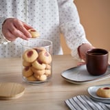 Person picks a cookie from a jar on a table, beside a cup of coffee and another cookie.