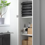 White ENHET high cabinet with stacked dark towels and toiletries on shelves.