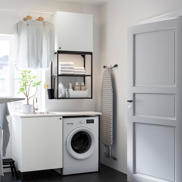 Compact white laundry setup with washing machine, sink, storage, and hanging space for clothes. Black accents highlight the organized design.