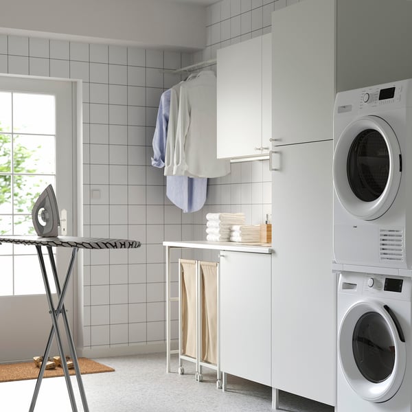 Modern laundry setup with white ENHET cabinets, stacked washer/dryer, clothes, ironing board, and storage.