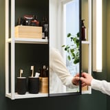 Image shows a person adjusting a bathroom mirror with shelves, holding toiletries and wooden boxes, reflecting a plant.