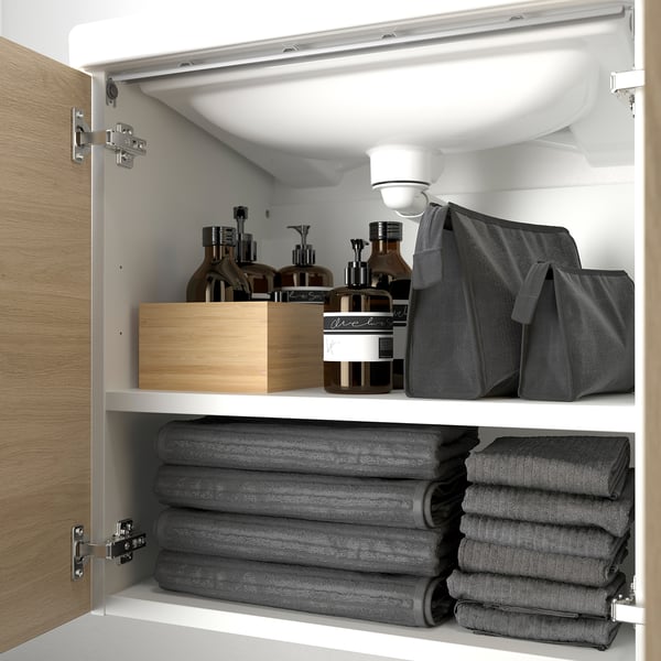 A well-organized bathroom cabinet with a wooden box containing toiletries on the top shelf and neatly folded gray towels on the bottom shelf.