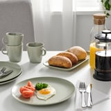 Table set with light green plates, holding food, mugs, and utensils. French press and carafe with juice are present.