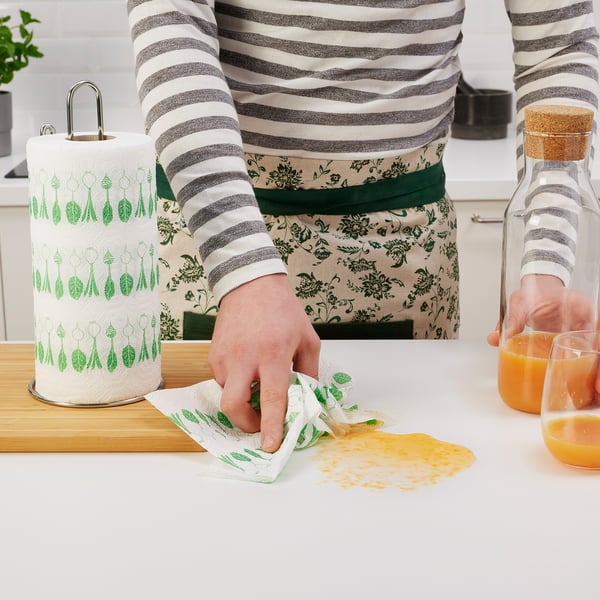 Woman cleans counter with absorbent, recycled FAMILJ kitchen roll featuring green vegetable patterns. Spilled juice nearby, apron worn.
