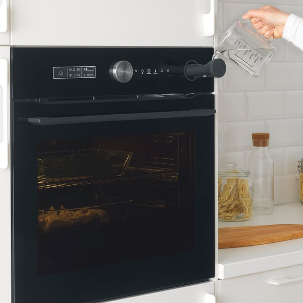 A hand pours water into a compartment on a modern black oven with digital controls and racks inside.