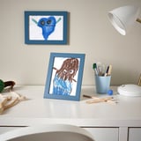 Childs desk with two drawings: blue monster, brown-haired figure. Art supplies, lamp.