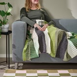 Woman relaxes on grey couch under green-striped blanket, holding metal cup. Plant and rug nearby.