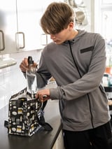 Person in gray jacket packs clear bottle into black FLADDRIG lunch bag with kitchen tools pattern in white kitchen.