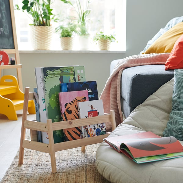 Bright living room with kids wooden bookrack, colorful books, grey couch, yellow chair, plants, and woven rug.