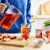Person pouring red tea with strawberries. FLÖJT tea box, plant, strawberries, ginger on table.