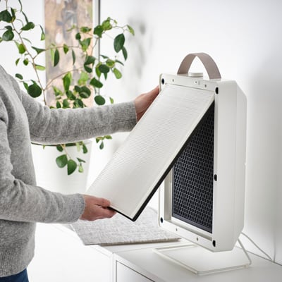 A person installs a white filter into a modern air purifier placed on a white stand. The filter helps remove particles like dust and pollen.