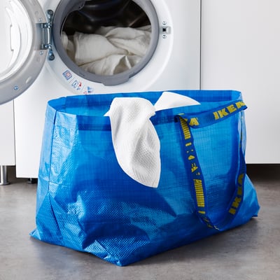 Blue ikea FRAKTA bag with white handles sits near a white washing machine, filled with white laundry.