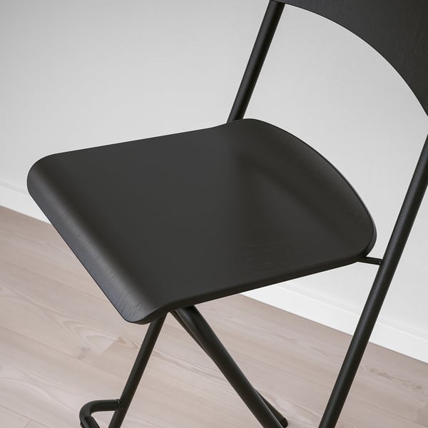 Black modern bar stool with curved seat and back, sturdy metal legs, shown against a light grey wall.
