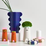 Whimsical table decor: blue wavy vase, orange face vases, white happy vase with greenery, colorful cylinders. Stoneware.