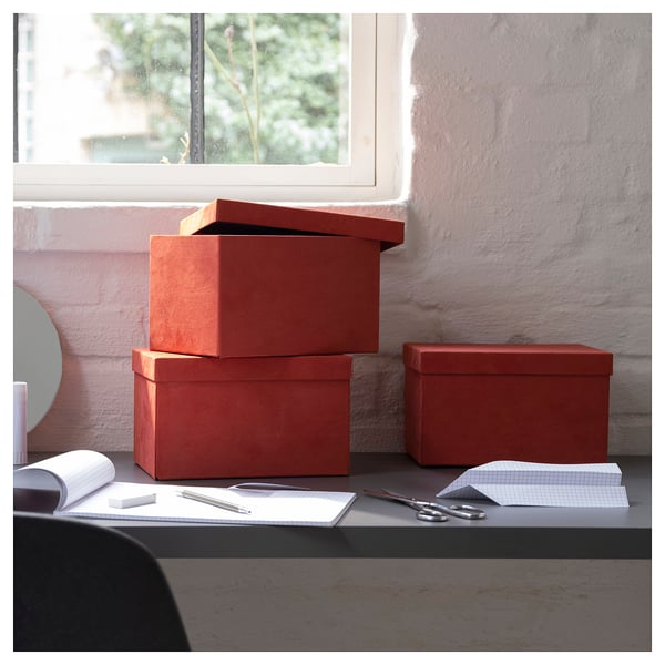 Three velvet red GJÄTTA boxes, stacked neatly, with an open notebook and scissors on a black desk by a window.