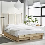 Minimalist bed: light wood frame, white linens, storage boxes, modern room, large windows, plants.