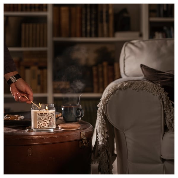 A person lights a GLANSLIND candle on a wooden table, next to a cozy chair with a blanket, in a warm room with bookshelves.