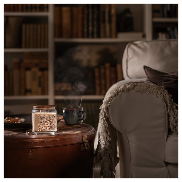 Cozy home scene with beige armchair, lit candle, and coffee mug on wooden side table. Behind, a bookshelf filled with books.