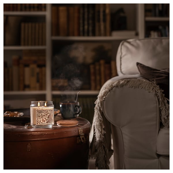 Cozy living room scene: lit scented candle, armchair with blanket, coffee mug, library background.