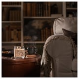 Cozy living room scene: lit scented candle, armchair with blanket, coffee mug, library background.