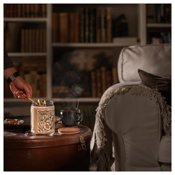 Hand lighting GLANSLIND candle in cozy room. Candle is white with brown label, on wooden table near cream armchair.