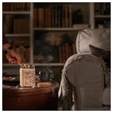 Hand lighting GLANSLIND candle in cozy room. Candle is white with brown label, on wooden table near cream armchair.