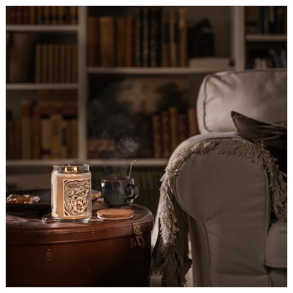 Cozy room setup: lit beige GLANSLIND candle on wooden side table beside armchair, blue mug, blanket.