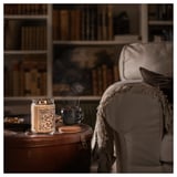 Cozy room setup: lit beige GLANSLIND candle on wooden side table beside armchair, blue mug, blanket.