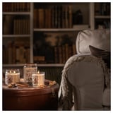 Cozy living room with lit candles on wooden trunk, armchair with blanket, bookshelf in background.