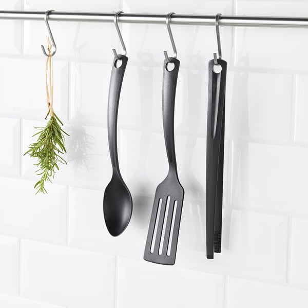 Three black kitchen utensils - spoon, spatula, tongs - hang from hooks. Herbs hang beside them on white wall.
