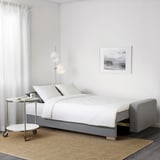 Minimalist bedroom with gray sofa-bed, white sheets, side table, lamp, and beach photo frame.