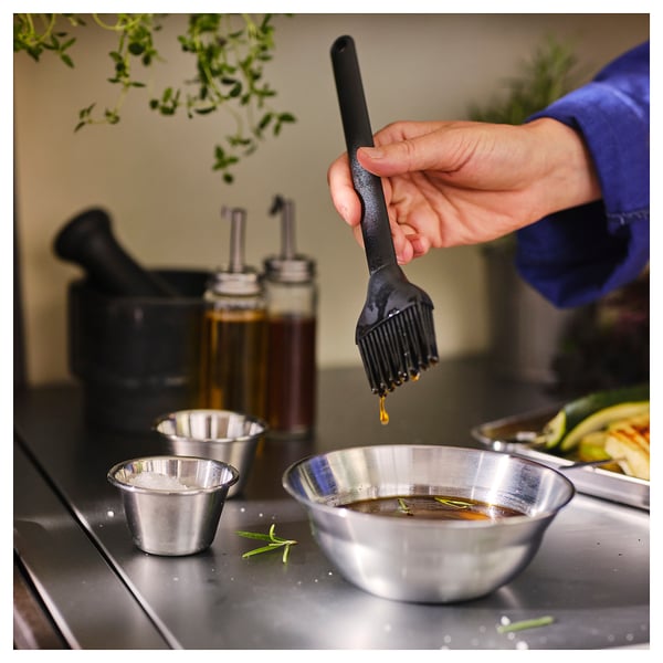 Person holding brush dipping into bowl of sauce, surrounded by metallic containers and herbs.