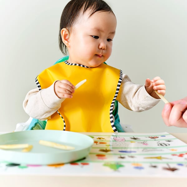 Baby wearing yellow bib, holds spoon, adult hand offers another. Table has colorful sticky notes.