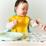 Baby wearing yellow bib, holds spoon, adult hand offers another. Table has colorful sticky notes.