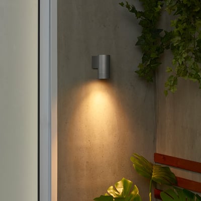 Small black GRÖNSPRÖT wall lamp emits warm light beside white door, surrounded by green plants, highlighting its compact size and sleek design.