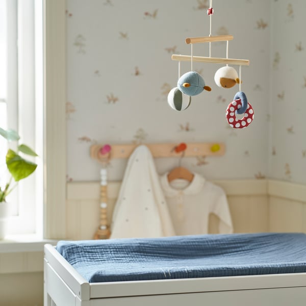 Baby mobile over changing table with toys. Nursery has floral wallpaper, coat rack, and blue pad.