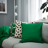 Cozy living room with grey sofa, green cushions, plant shelf, and large window.