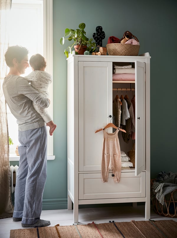 Man with baby by green HÄNGA wardrobe, childs clothes, plant on top.