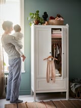 Man with baby by green HÄNGA wardrobe, childs clothes, plant on top.