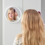 A girl brushes her blonde hair seen in a circular mirror attached to a white wall.