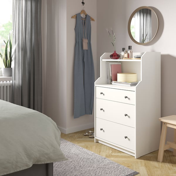 Modern bedroom with white HAUGA chest, mirror, blue jumpsuit, plants, & minimalist decor.