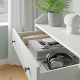 White HAUGA chest with open drawer showing books, headphones, and cables. Modern, minimalist design with a top shelf and safety fitting.