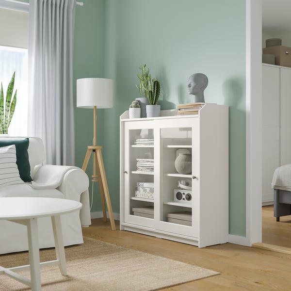 Bright modern living room with mint walls, white furniture, and natural light. Centered is a white HAUGA cabinet with glass doors.