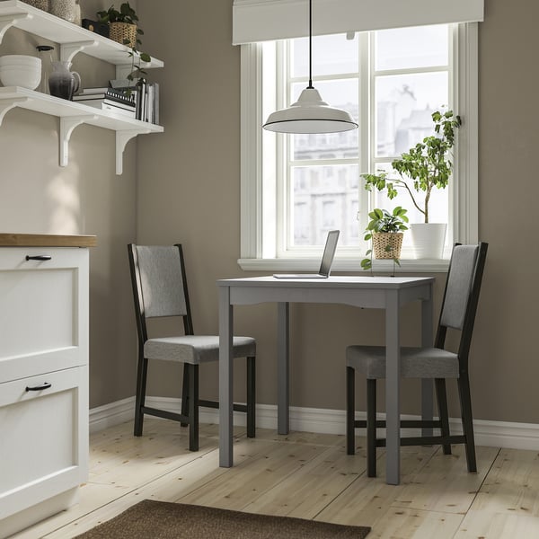 Compact dining set with gray table & matching chairs near window, white shelves, light wood floors.