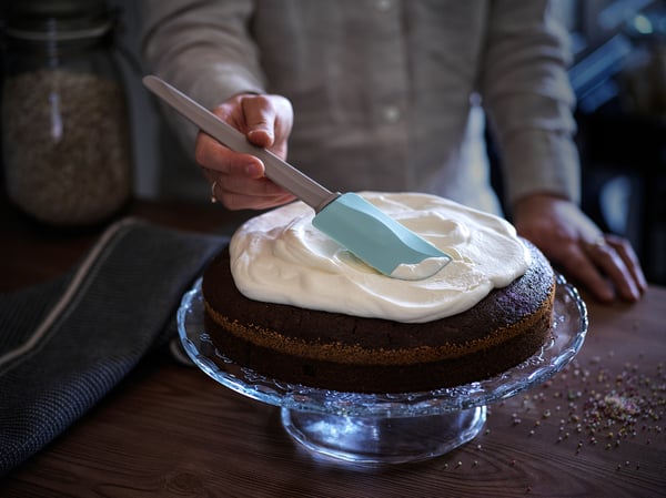 Frosting cake on glass stand with blue spatula. Two-layered cake with white frosting on wooden table.