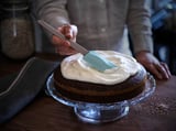 Frosting cake on glass stand with blue spatula. Two-layered cake with white frosting on wooden table.