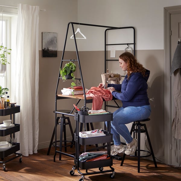 A woman sews at a black table with a storage ladder, surrounded by black trolleys and a stool, with plants and art in the background.